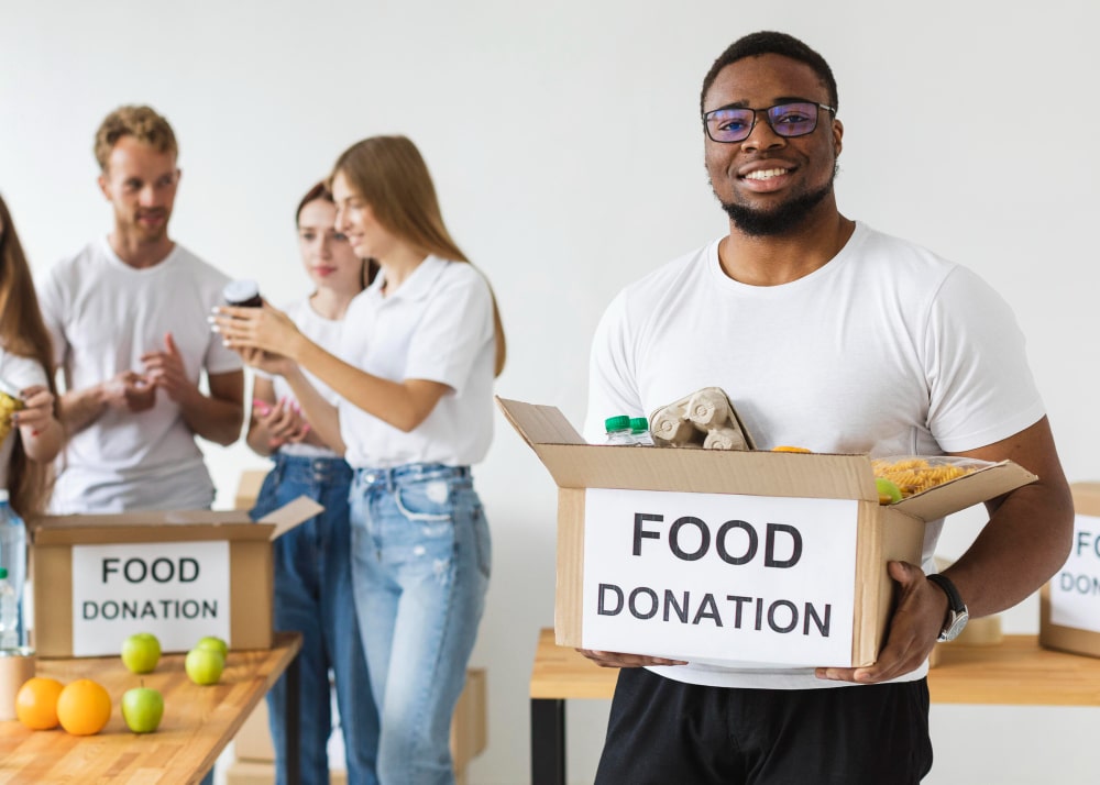 A smiling person holds a box labeled "Food Donation," filled with groceries. Behind, three people pack items.