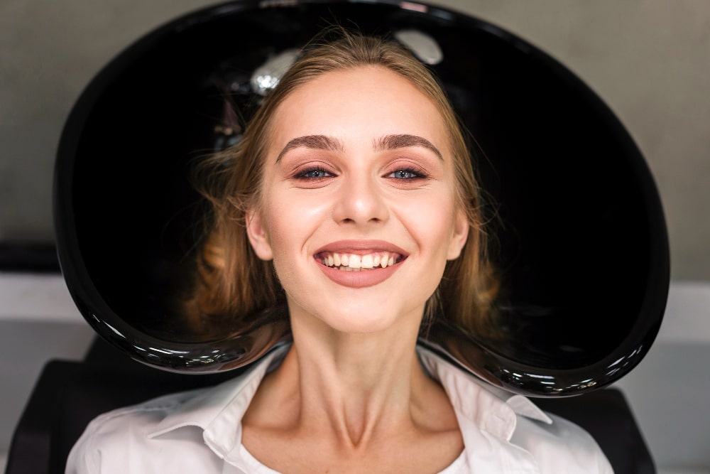 A young woman smiles in a salon, reclining at a black wash basin with a radiant glow.