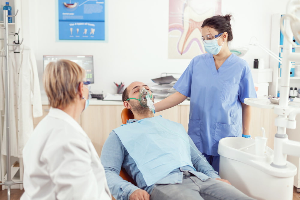 A male patient in a dental clinic wearing a mask, attended by two female dentists.