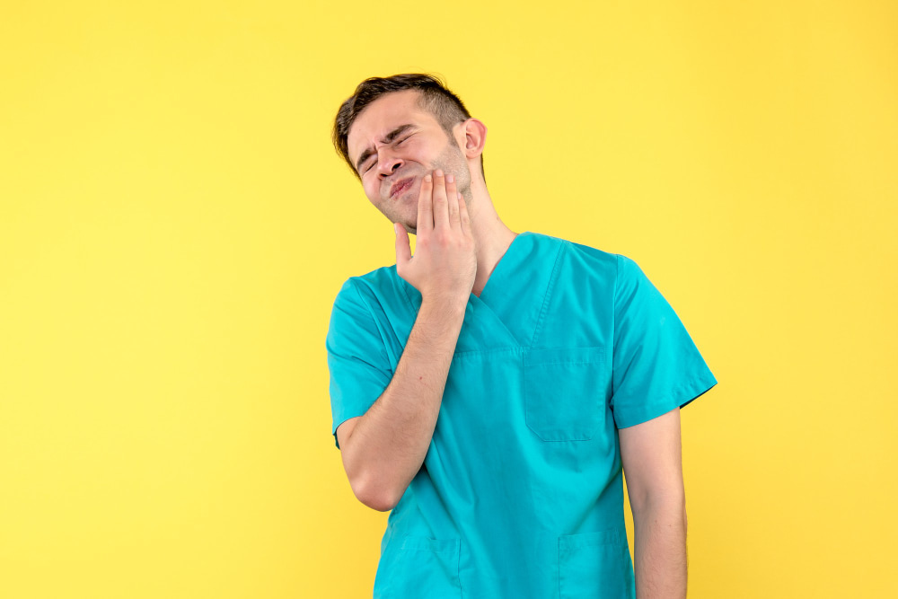 A man wearing blue scrubs is touching his cheek with a pained expression.