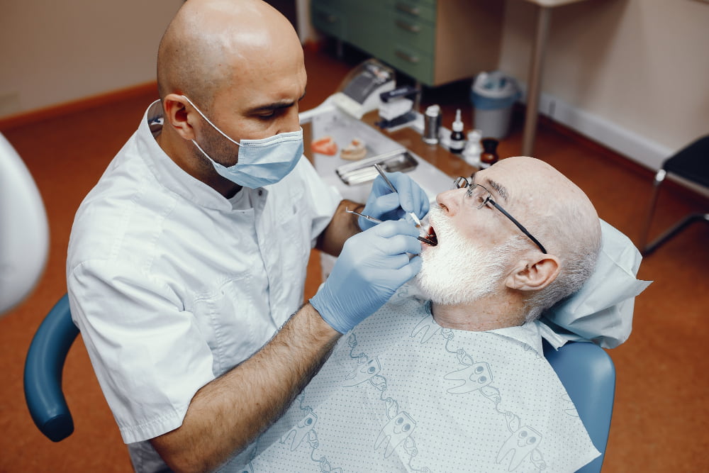 A dentist in a white coat and mask examines an elderly man with a white beard in a dental chair.