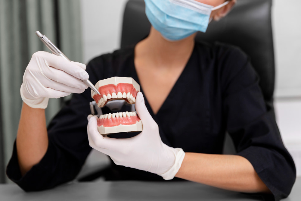 A masked dentist in black scrubs demonstrates dental care using a set of dentures and holding a dental tool.