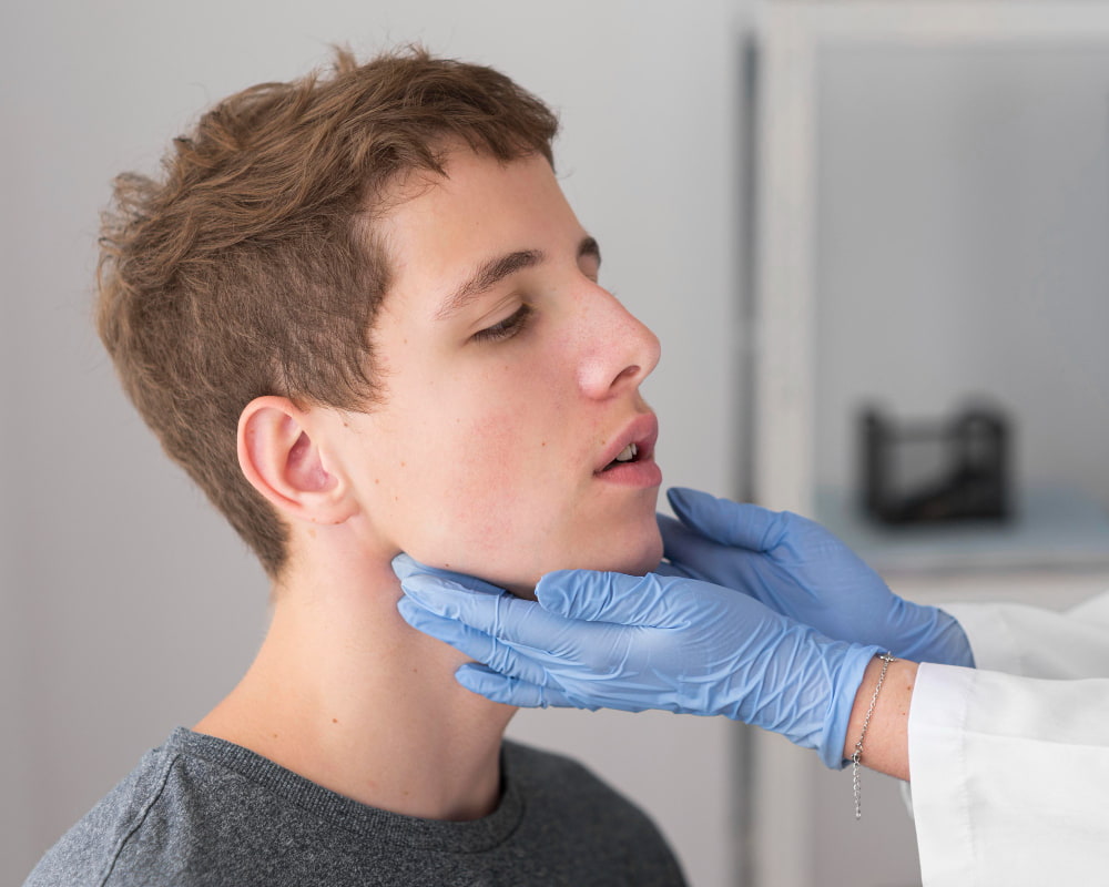 A doctor in blue gloves gently examines a patient's jawline as they sit sideways with eyes closed in a clinical setting.